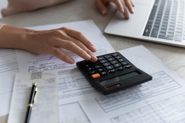 Crop close up of female sit at desk calculate expenses expenditures on machine pay bills taxes online on computer. Woman work with financial documents manage family budget. Bank, finances concept.